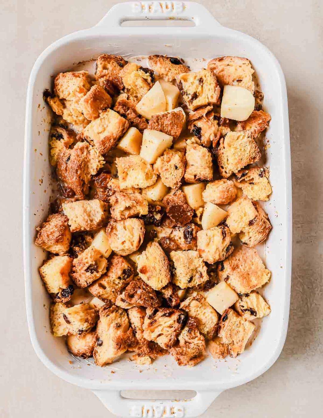 A white baking dish filled with golden-brown bread pudding, featuring chunks of bread and apples, lightly browned and moist.