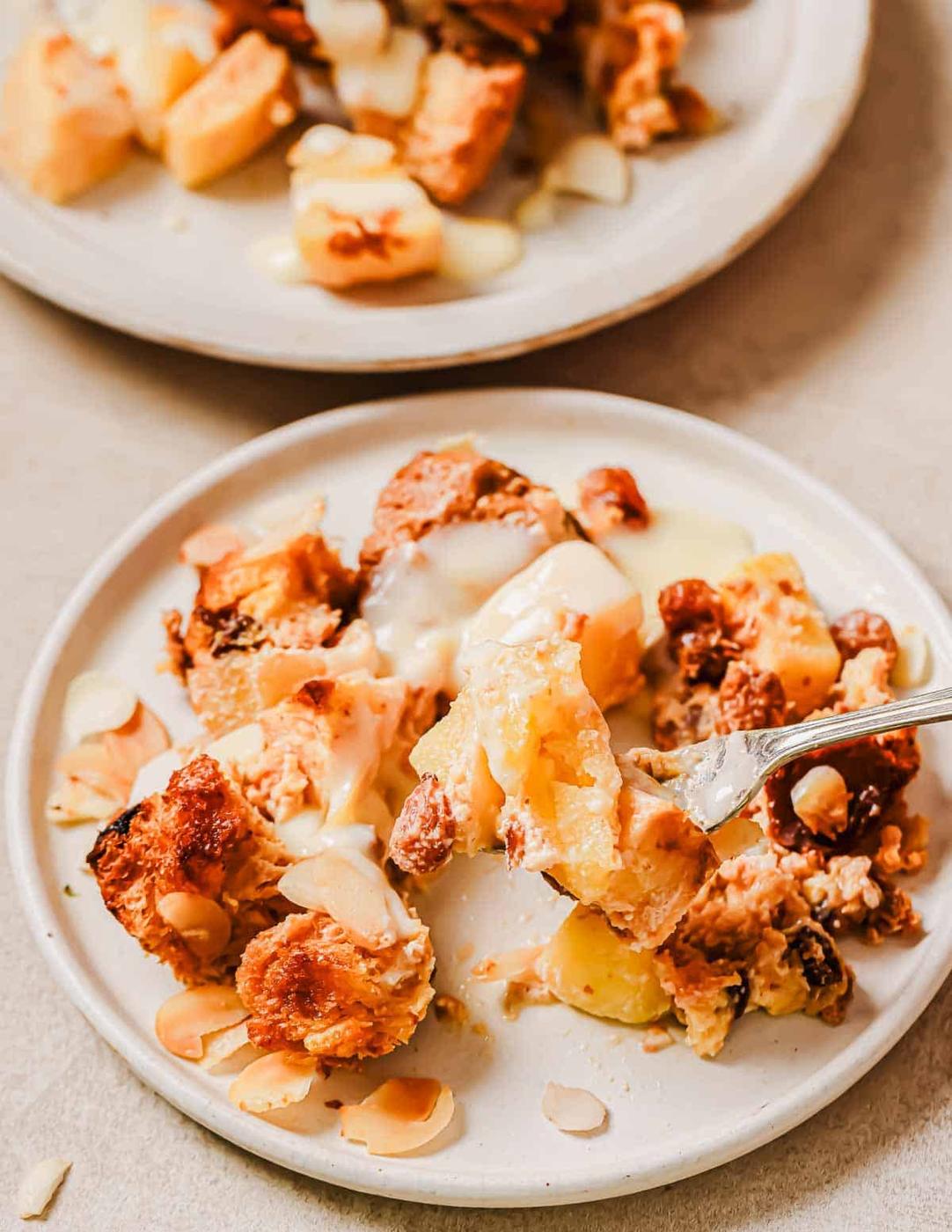 A plate of bread pudding topped with sliced almonds and drizzled with custard. In the background, there's another plate with a similar dessert. A fork rests on the front plate, holding a bite-sized piece.