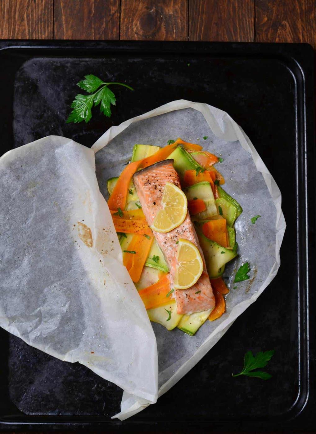 A piece of salmon baked in parchment paper with vegetables. The vegetables appear to be carrots and zucchini. Lemon slices are placed on top of the salmon. The parchment paper packet sits on a dark baking sheet. Small sprigs of parsley are on the baking sheet near the packet. The background is a dark wooden surface.