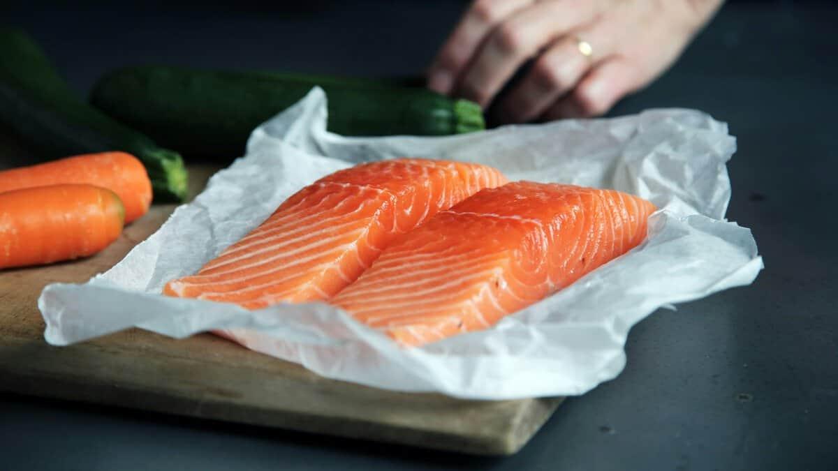 Two salmon fillets are neatly arranged on parchment paper atop a wooden cutting board. Baby carrots and zucchini are visible in the background, slightly out of focus. A hand is partially visible in the upper right corner.
