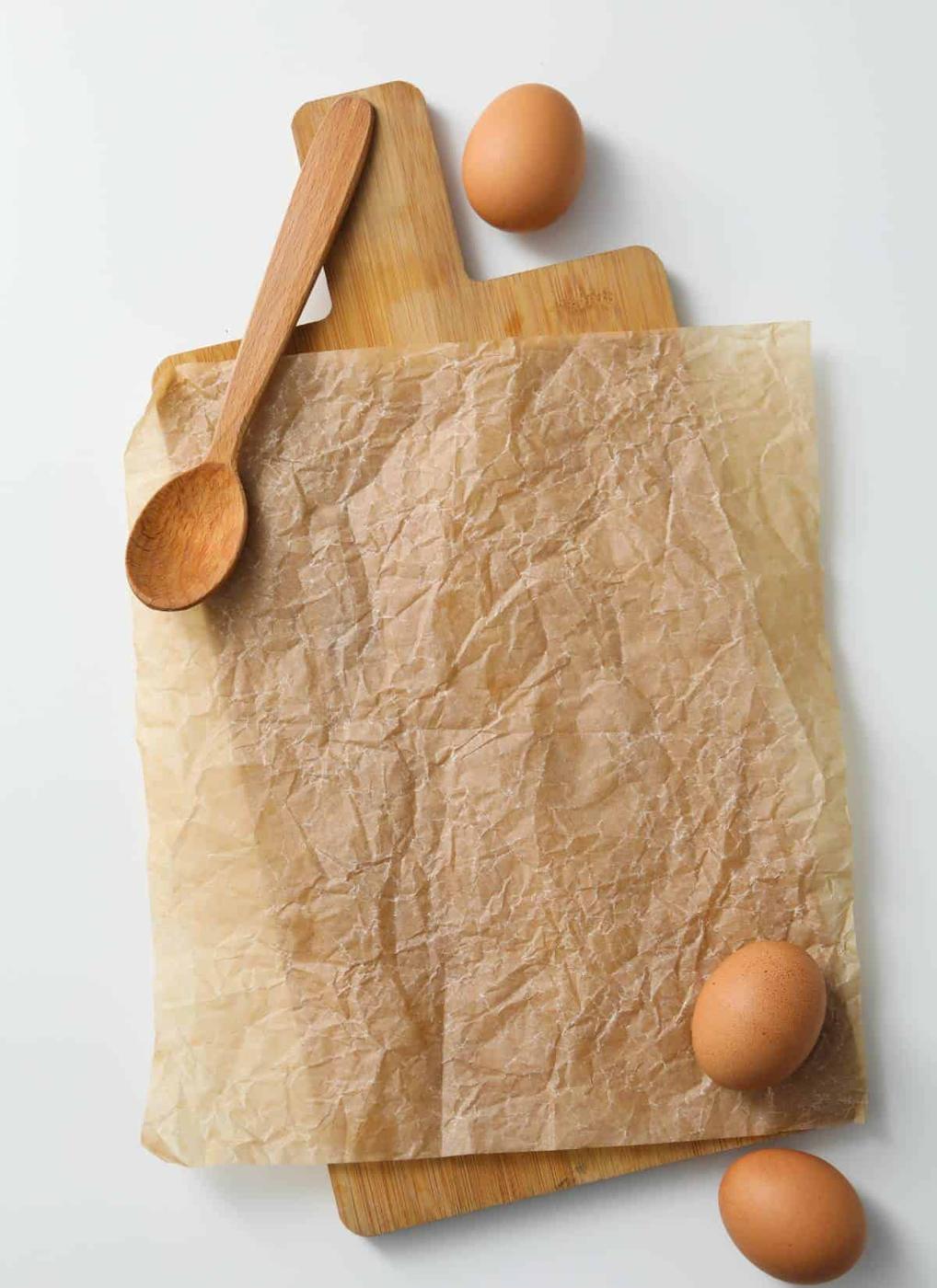 A flat lay image of a wooden cutting board with crumpled brown parchment paper on top. A wooden spoon and three brown eggs are arranged around the board.