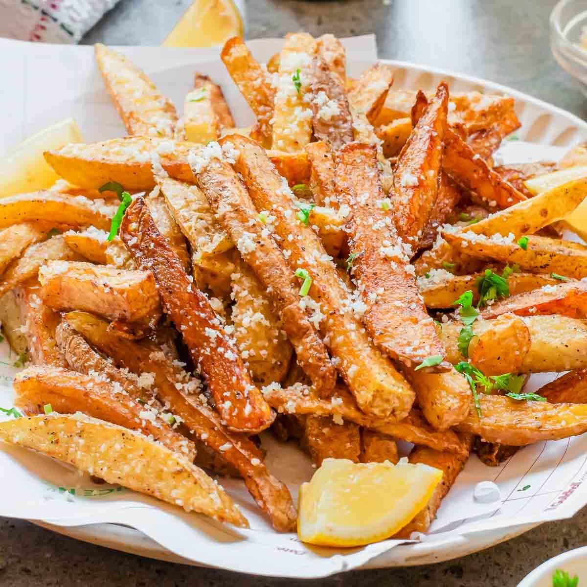 A plate of crispy parmesan truffle fries topped with chopped herbs, served with lemon wedges on the side.