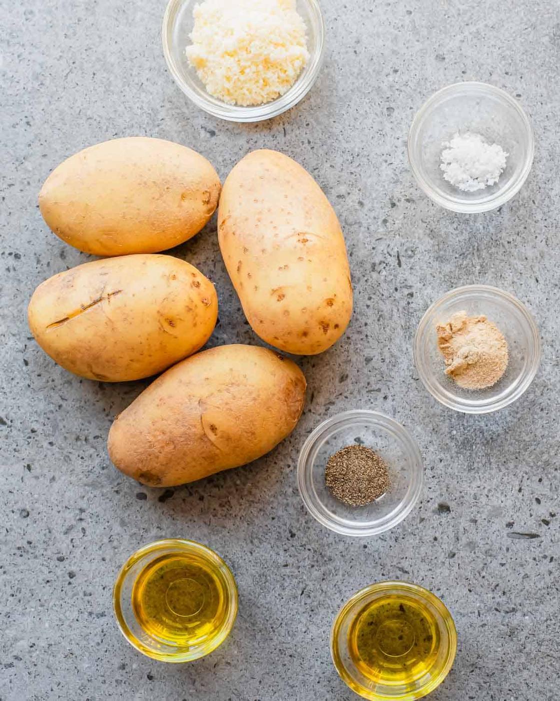 Four whole potatoes surrounded by small glass bowls containing grated cheese, salt, ground pepper, garlic powder, and two bowls of oil, all arranged on a gray countertop.
