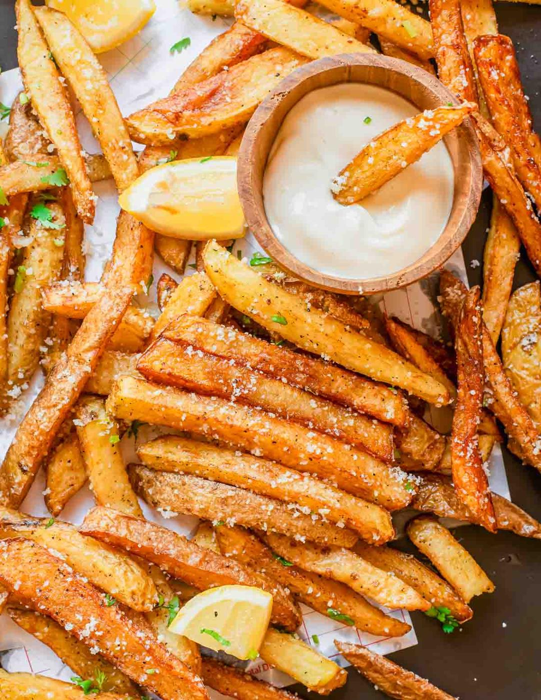 A pile of golden, crispy truffle French fries topped with grated parmesan cheese and parsley, served with lemon wedges and a bowl of creamy dipping sauce. One fry is dipped in the sauce.