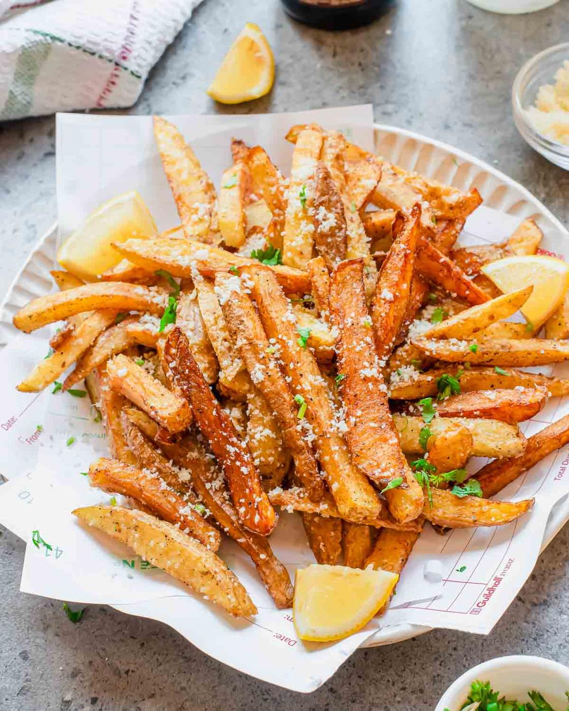 A plate of golden, crispy truffle French fries topped with grated cheese and chopped herbs, garnished with lemon wedges, served on parchment paper.