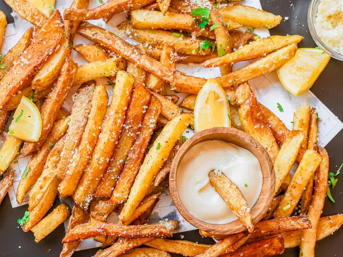 A pile of golden truffle fries sprinkled with herbs and grated parmesan cheese, garnished with lemon wedges. A small wooden bowl of creamy dipping sauce sits among the fries, with one fry dipped inside.