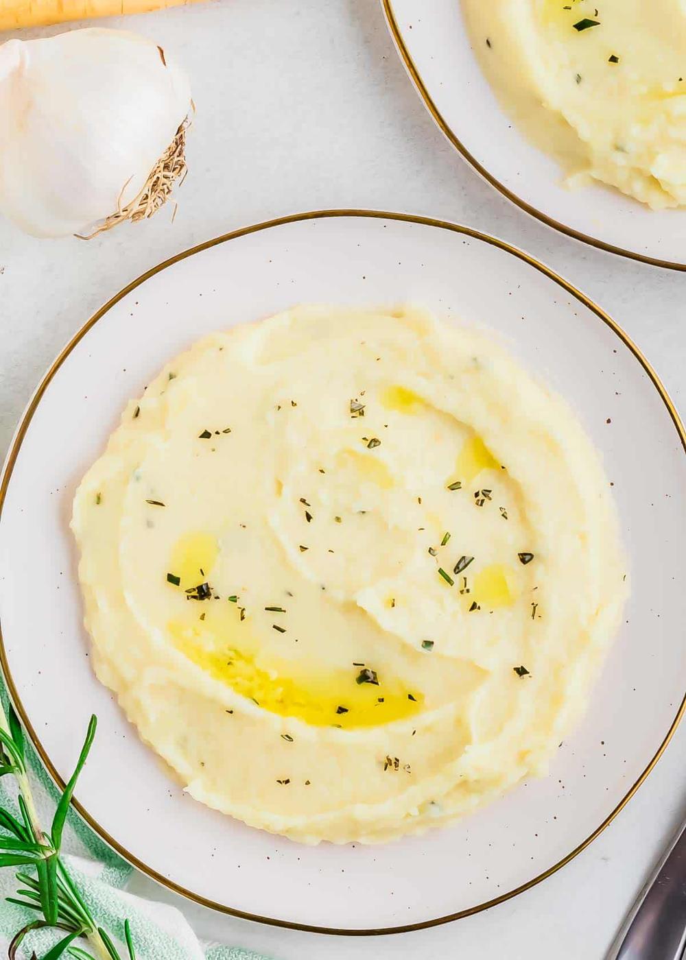 Plate of creamy mashed parsnips garnished with herbs, with garlic and rosemary in the background.