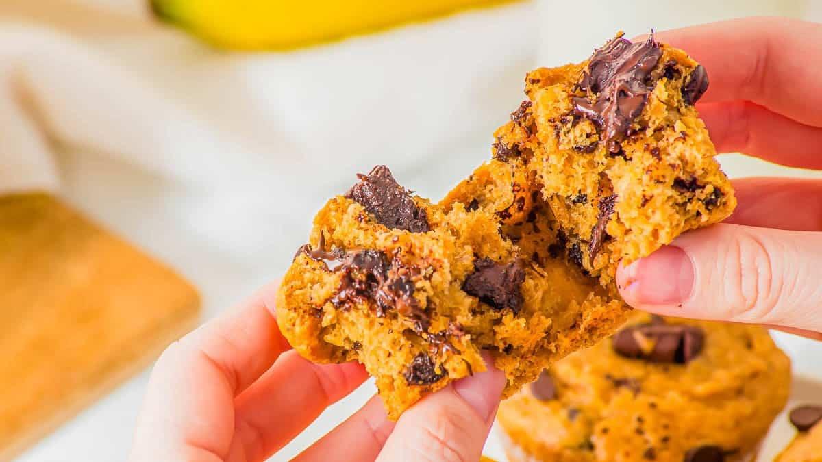 Someone's hands are breaking apart a freshly baked muffin filled with melted chocolate chunks. The muffin appears moist and rich, with a golden-brown color. A blurred background shows more muffins and a hint of a banana.
