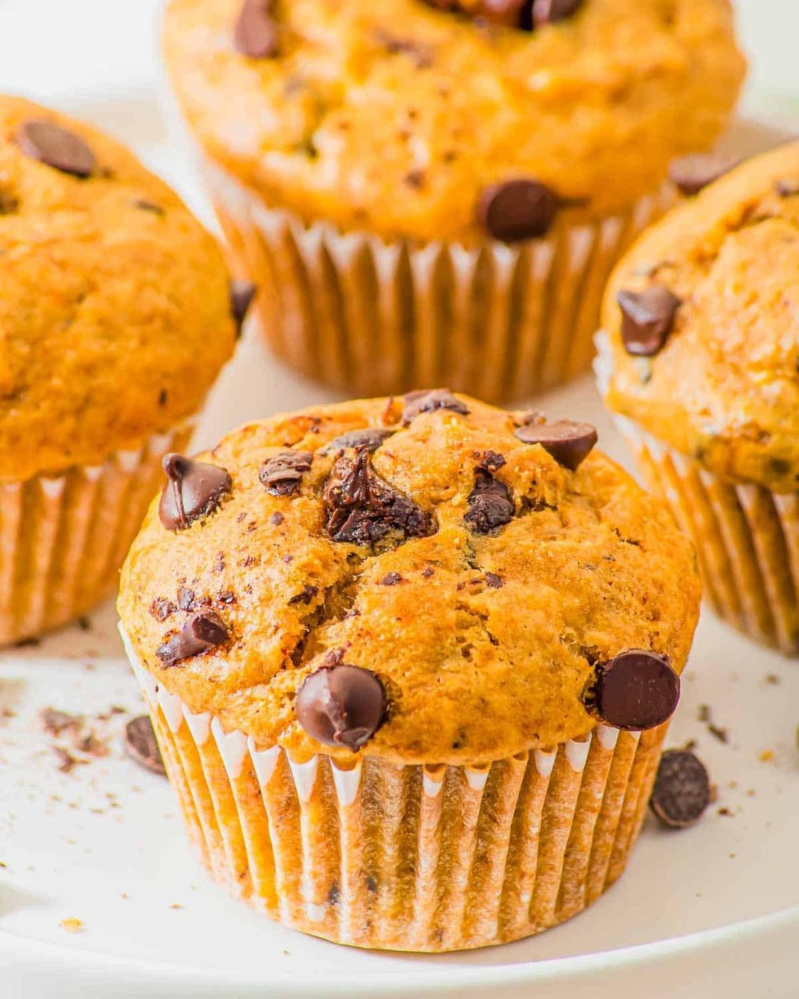 Four freshly baked muffins with chocolate chips on a white plate. The muffins have a golden-brown color and are topped with scattered chocolate chips. Some crumbs and chips are visible on the plate.
