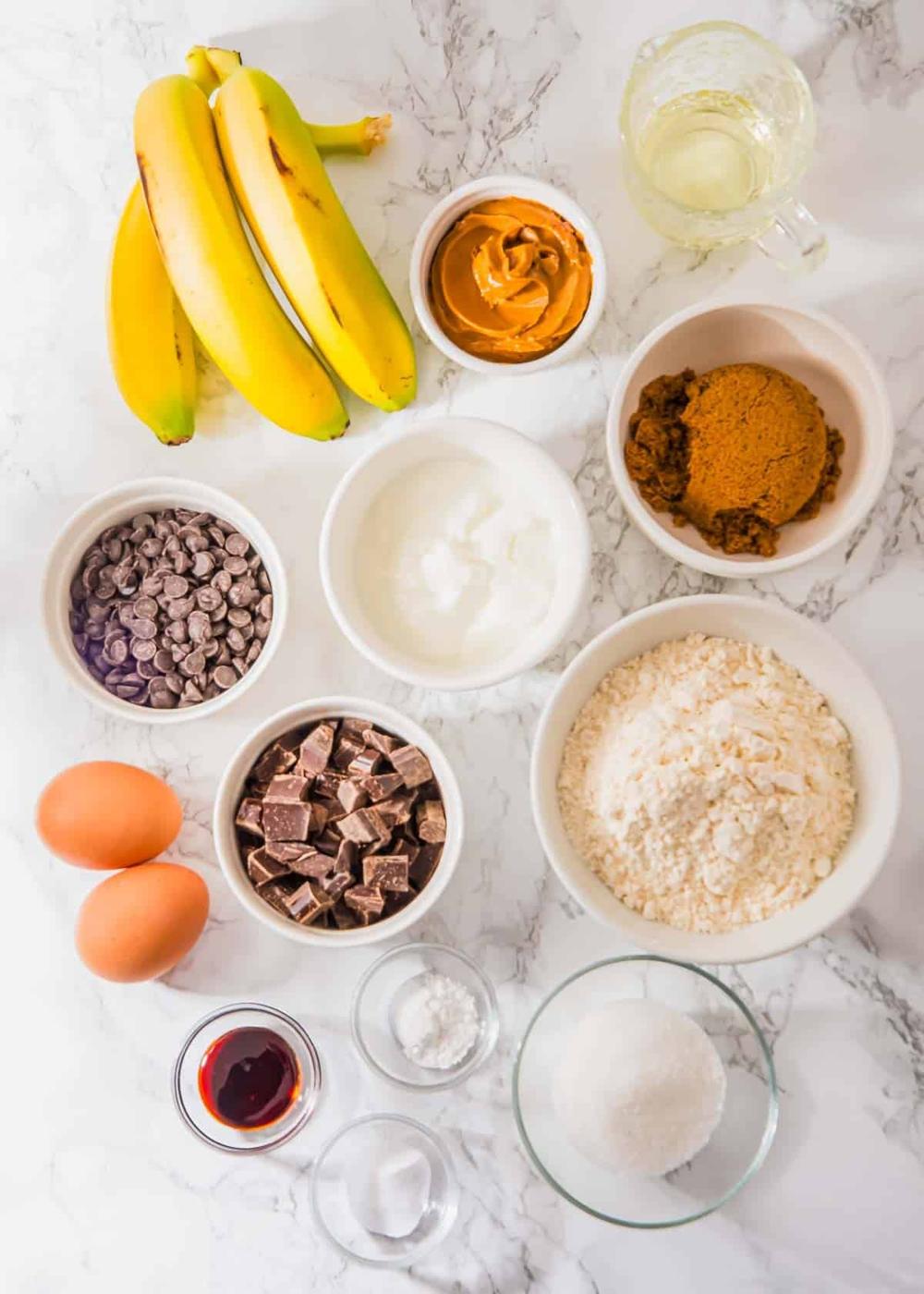 Bowls of chocolate chips, yogurt, flour, brown sugar, and peanut butter, along with whole bananas, eggs, vanilla extract, baking soda, and oil, are arranged on a white marble surface.
