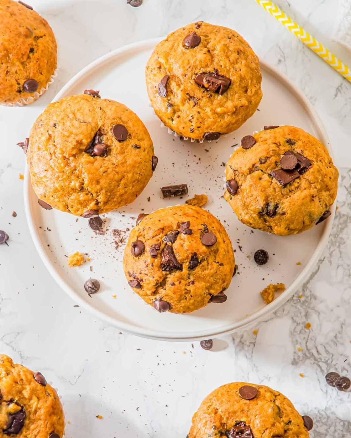 A plate of six banana peanut butter muffins on a marble surface. Some chocolate chips and muffin crumbs are scattered around, with a yellow and white straw partially visible at the edge.