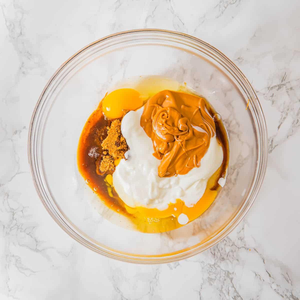 A glass bowl contains a mixture of creamy peanut butter, a cracked egg, yogurt, brown sugar, and vanilla extract. The ingredients, not yet mixed, rest on a white marble surface.