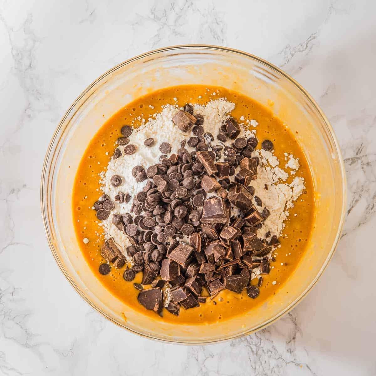 A mixing bowl containing a peanut butter banana muffin batter with flour, chocolate chips, and chocolate chunks on top, placed on a white marble surface.
