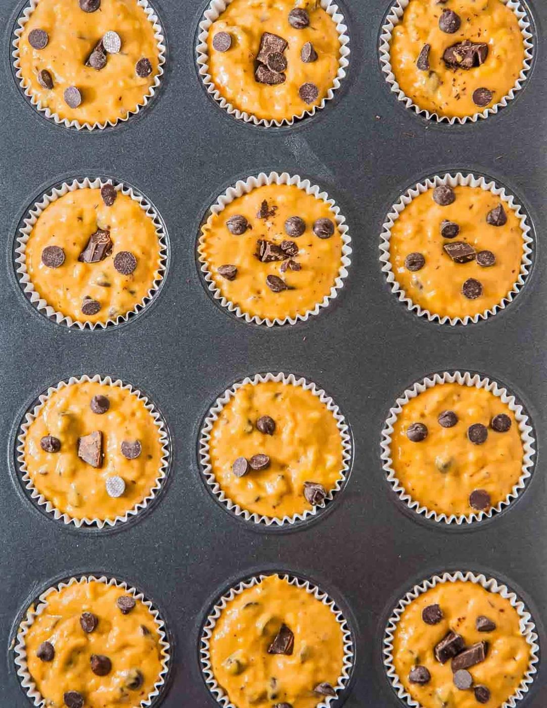 A muffin tray filled with unbaked peanut butter banana muffins in paper liners, each topped with chocolate chips and chunks. The batter is light brown, and the arrangement is in a grid with three rows of four muffins.