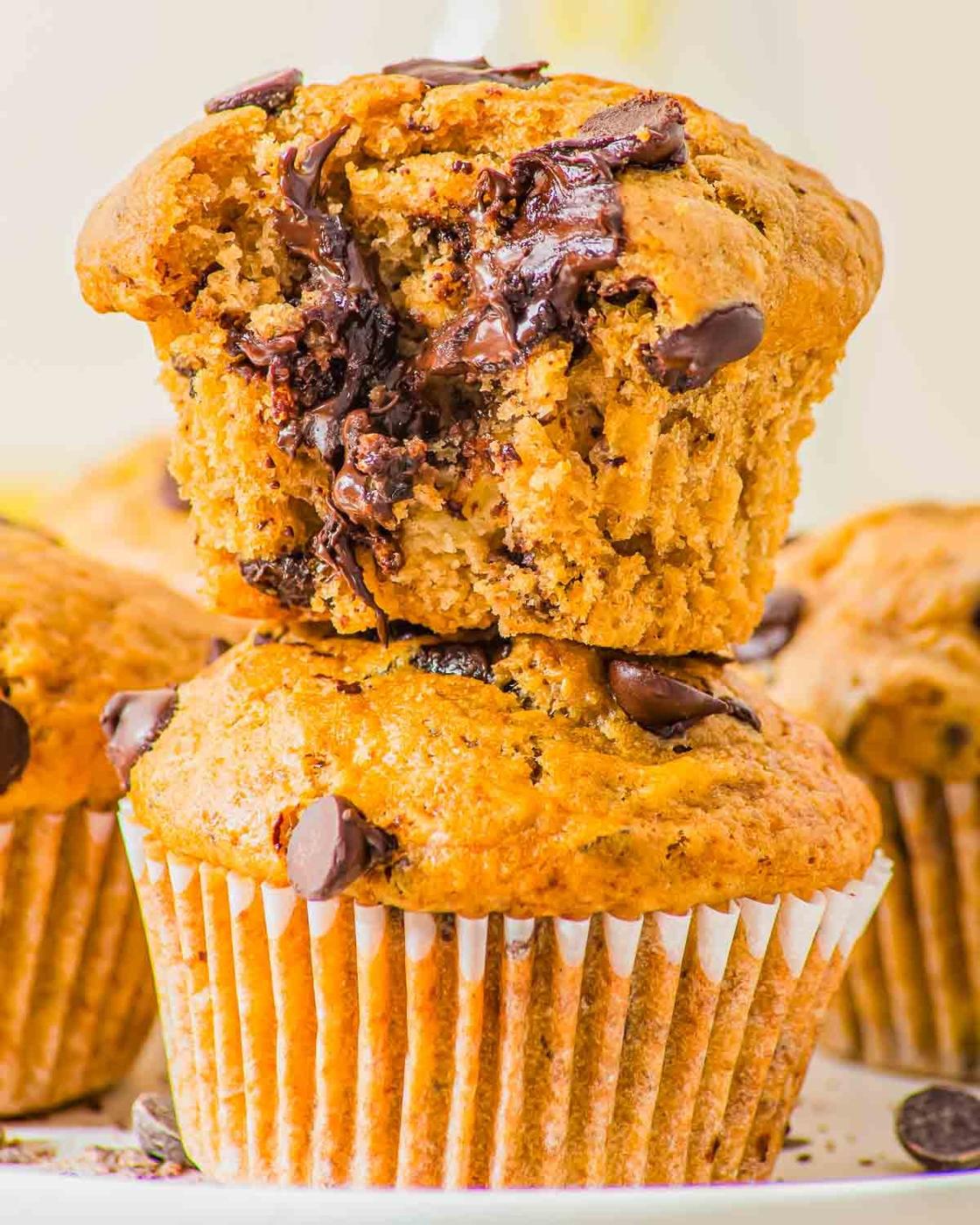 Two peanut butter banana muffins are stacked on a plate. The top muffin has a bite taken out of it, revealing a soft, moist interior with melted chocolate chips. The muffins have a golden-brown color and are in white paper liners.
