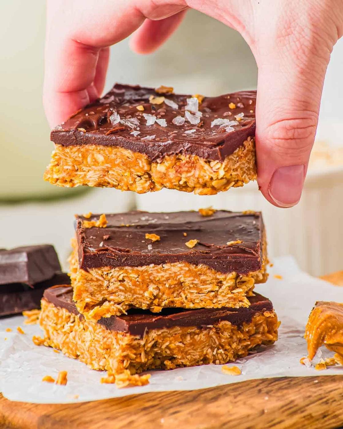 A hand lifts a chocolate-topped oat bar from a stack of three bars on parchment paper. The bars have a golden oat base and a thick, glossy chocolate layer sprinkled with flaky sea salt.
