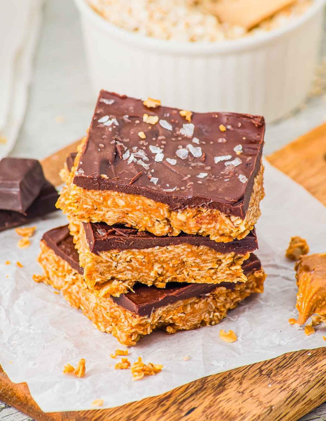 Three stacked chocolate-topped oat and peanut butter bars sit on parchment paper, sprinkled with flaky sea salt. More oat mixture, chocolate, and a bowl are blurred in the background on a wooden surface.