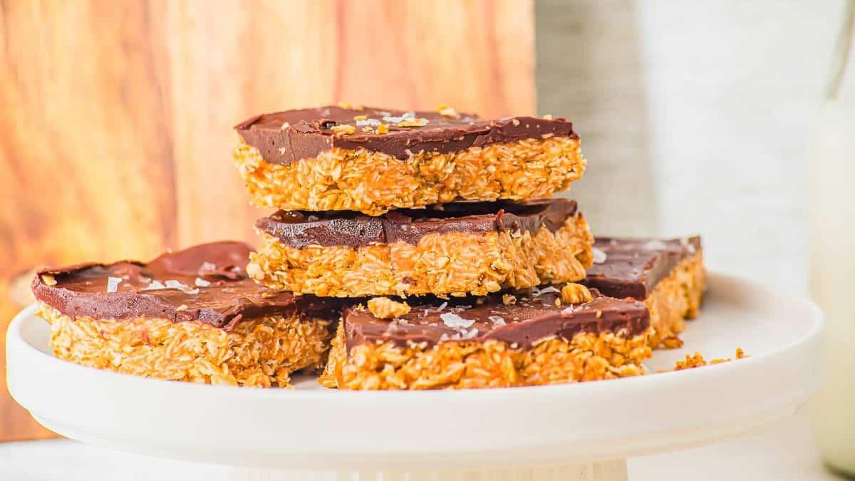 Five chocolate-topped oatmeal peanut butter bars stacked on a white cake stand, with a wooden board in the background. The bars have a thick oat layer and are sprinkled with flakes of sea salt.