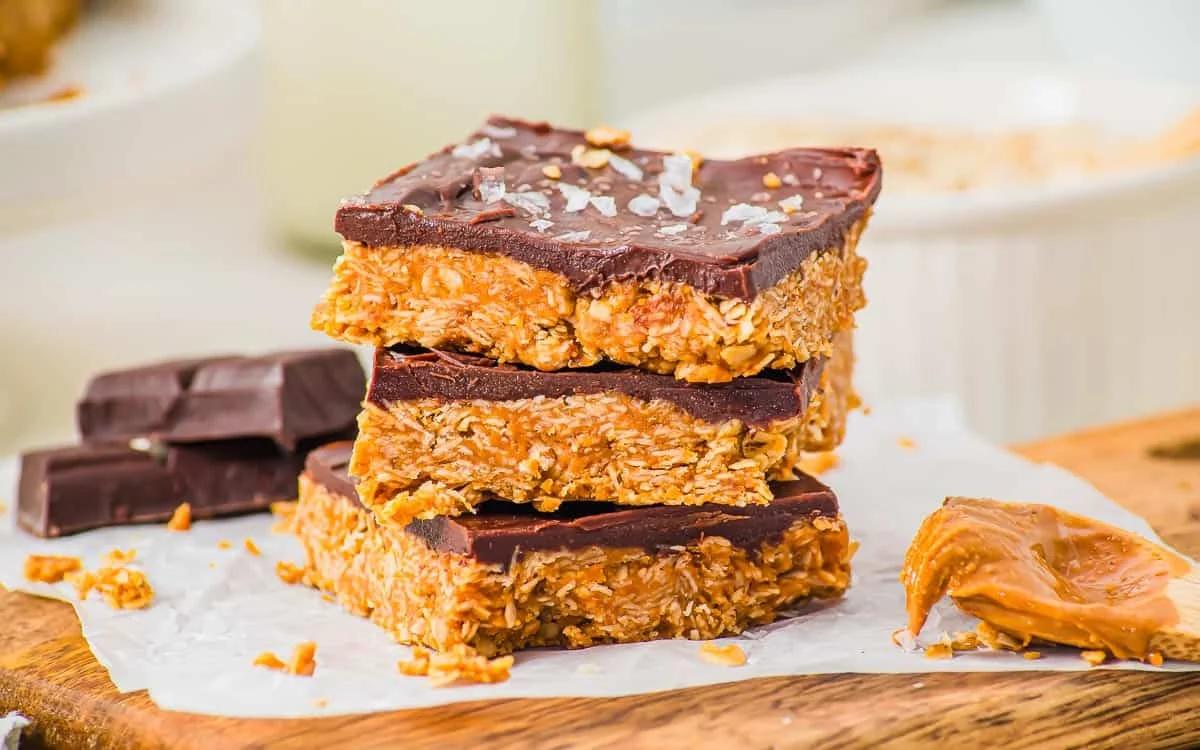 Three no-bake chocolate peanut butter oat bars stacked on parchment paper, topped with sea salt. A chunk of chocolate and a spoonful of peanut butter are nearby on a wooden surface.
