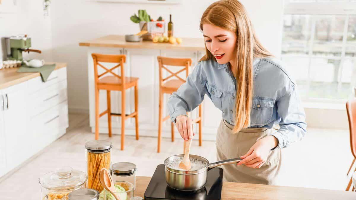 A woman stirs a pot on an induction stove in a bright kitchen. There are jars of pasta and spices on the counter, and she is wearing a blue shirt and apron, with her hair down.