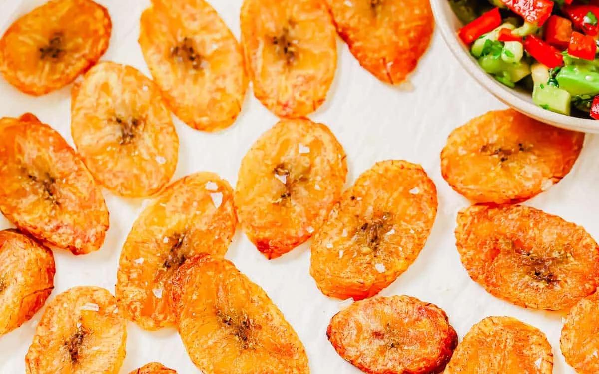 Golden, crispy baked plantain chips are arranged on a white surface next to a bowl of colorful chopped salsa.