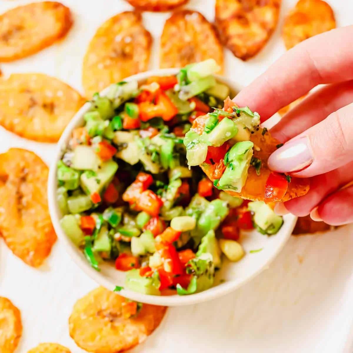 A hand holds a plantain chip topped with a colorful salsa made of chopped kiwi, avocado, red bell pepper, and herbs, with more chips and salsa in a bowl on a light surface.