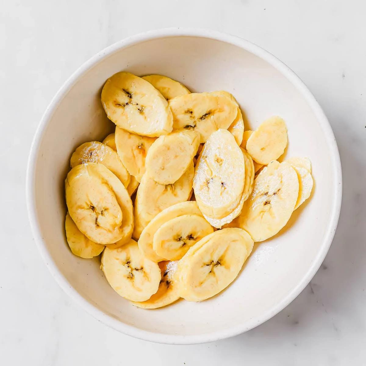 A white bowl filled with thin, round slices of raw plantain on a light marble surface.