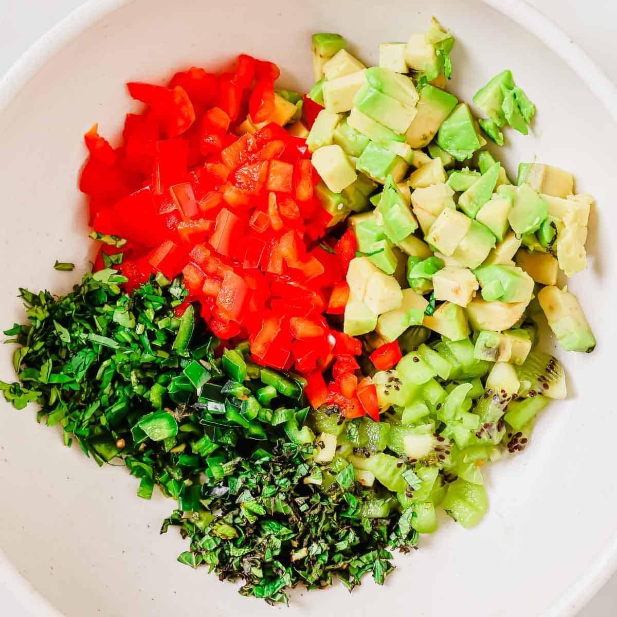 A bowl containing chopped red bell pepper, avocado, kiwi, jalapeño, and fresh herbs arranged in separate sections to make a salsa for serving with plantain chips.