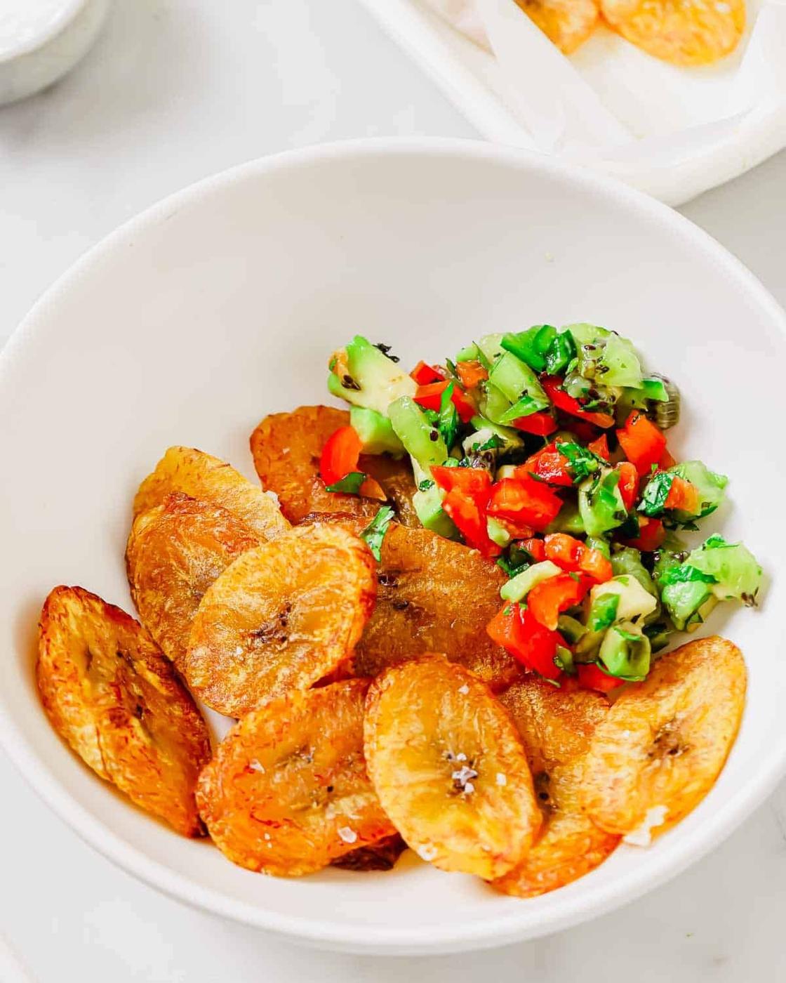 A white bowl containing fried plantain chips and a colorful avocado, red bell pepper, and kiwi salsa.
