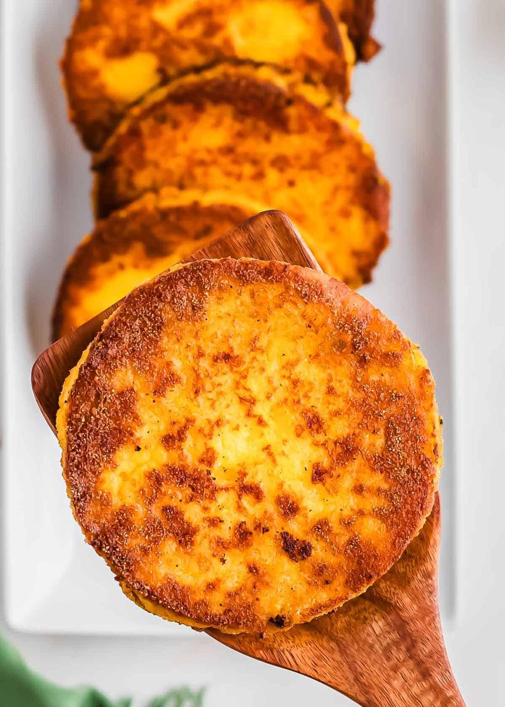 Close-up of a golden, crispy arepa on a wooden spatula, with more polenta cakes blurred in the background on a white plate. The texture shows a well-cooked, slightly browned surface.
