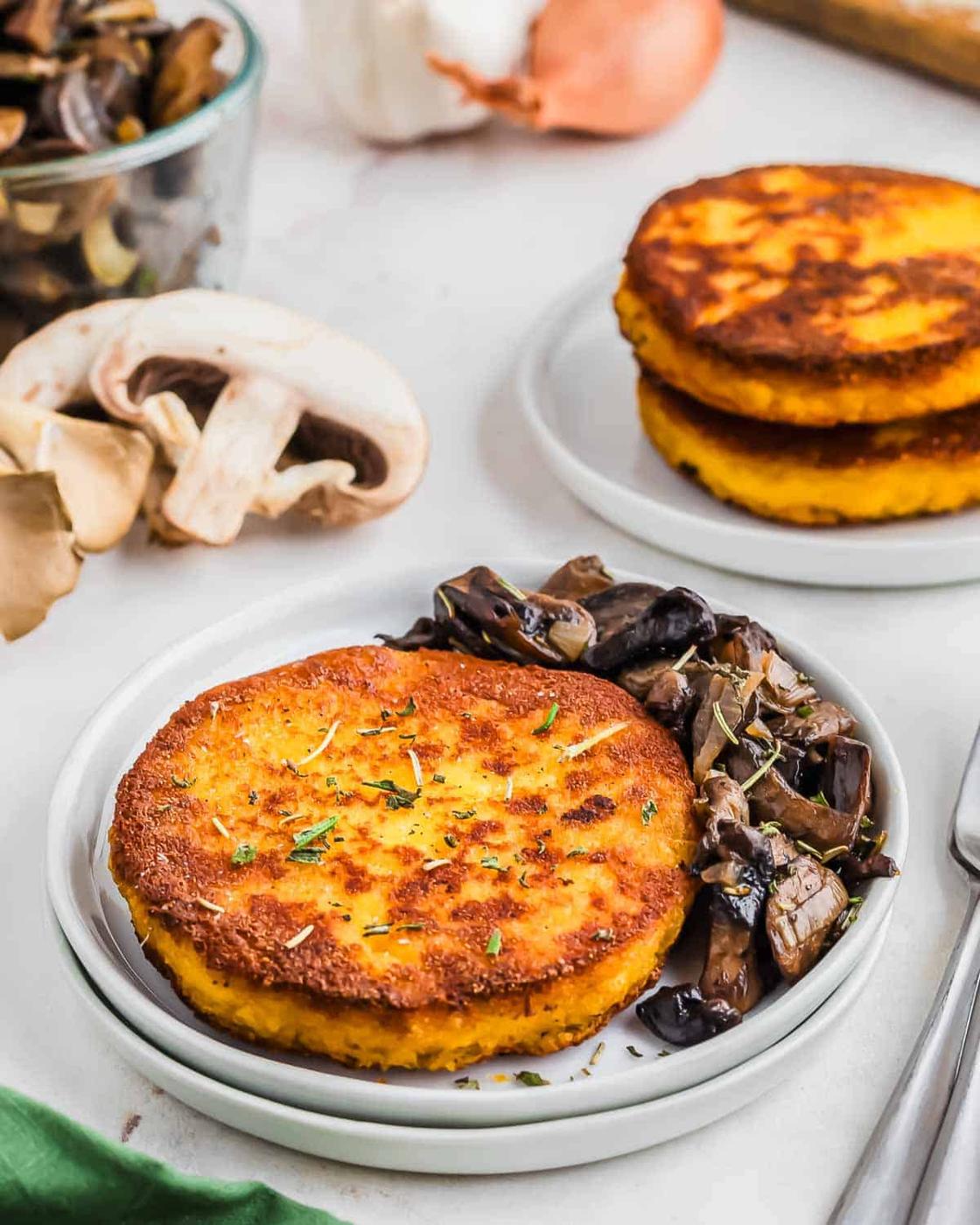 Two golden-brown polenta cakes garnished with herbs on a white plate, accompanied by sautéed mushrooms. A glass bowl with sliced mushrooms and a garlic bulb are in the background, adding a rustic touch to the setting.