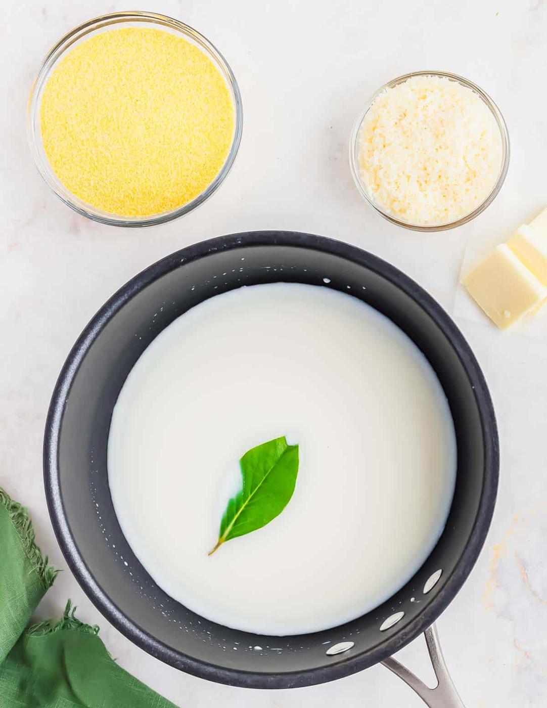 A black saucepan with milk and a single green bay leaf sits on a white surface. Nearby are two glass bowls with cornmeal and grated cheese, and two pieces of butter. A green cloth is partially visible at the bottom left.
