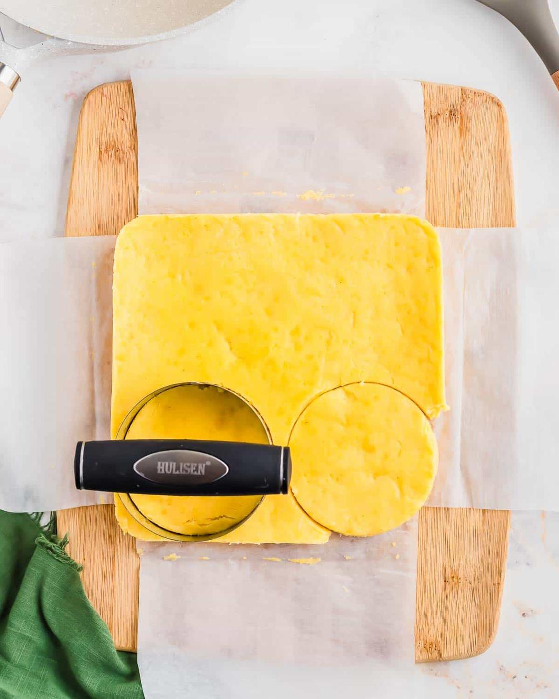 A square piece of cooked polenta on a wooden board, partially covered with parchment paper. A round cookie cutter is placed on the polenta, cutting out a circular shape. A green cloth is partially visible at the bottom left corner.