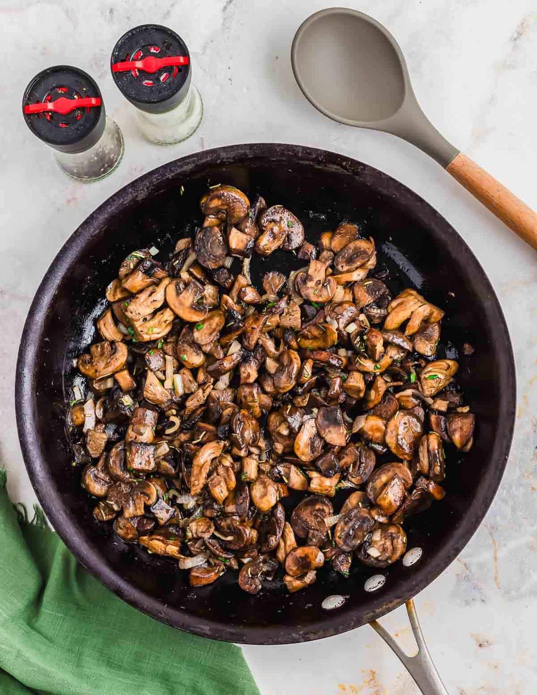 A skillet filled with cooked sliced mushrooms and herbs, placed on a light marble surface. A gray spoon with a wooden handle rests beside the skillet, and two shakers with red caps are nearby. A green cloth is partially visible in the corner.
