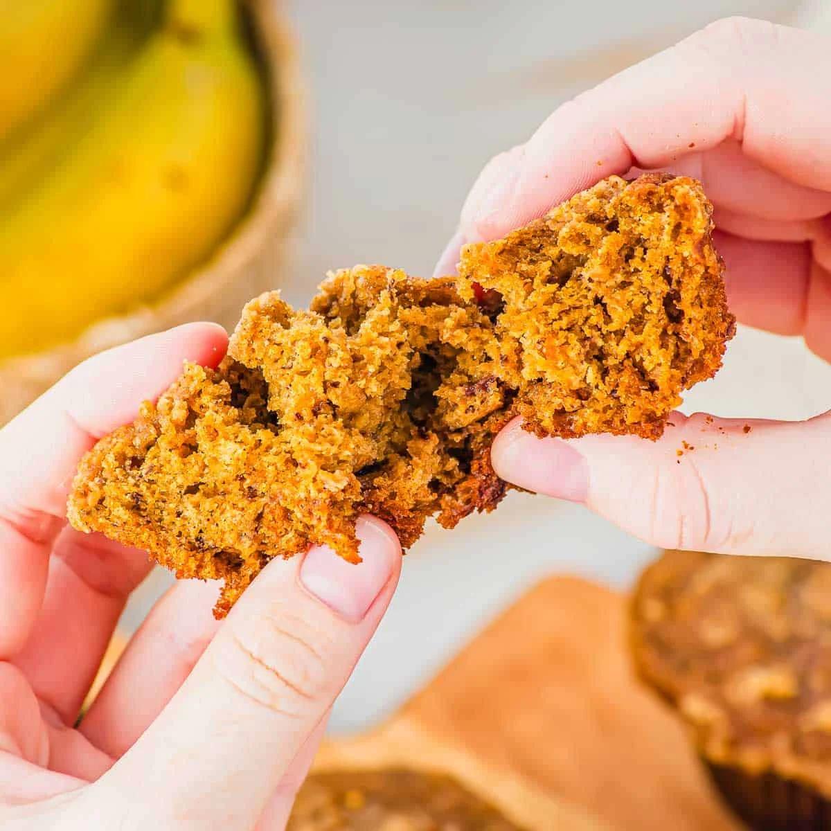 Close-up of hands breaking open a moist banana pumpkin muffin, with bananas in the background on a table.