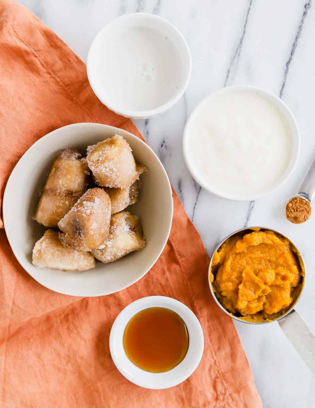 Frozen bananas, milk, yogurt, pumpkin, pumpkin pie spice and maple syrup in small bowls on a marble surface with an orange napkin.
