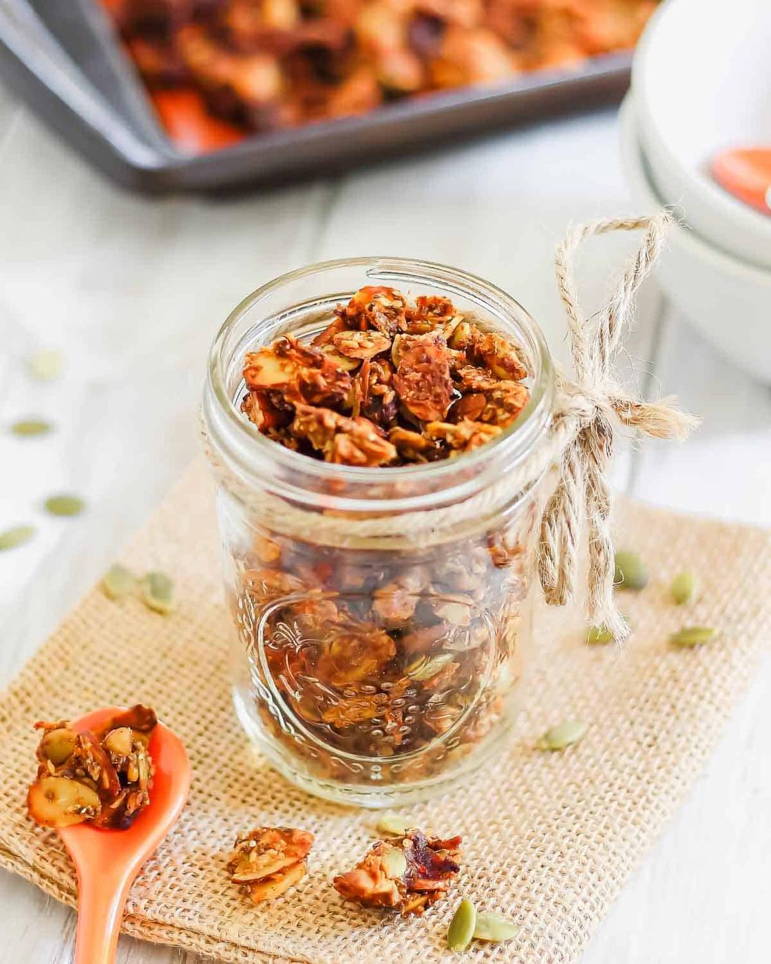 A mason jar filled with homemade pumpkin granola sits on a burlap cloth, with a small orange spoon containing more granola beside it. A tray of granola is in the background.