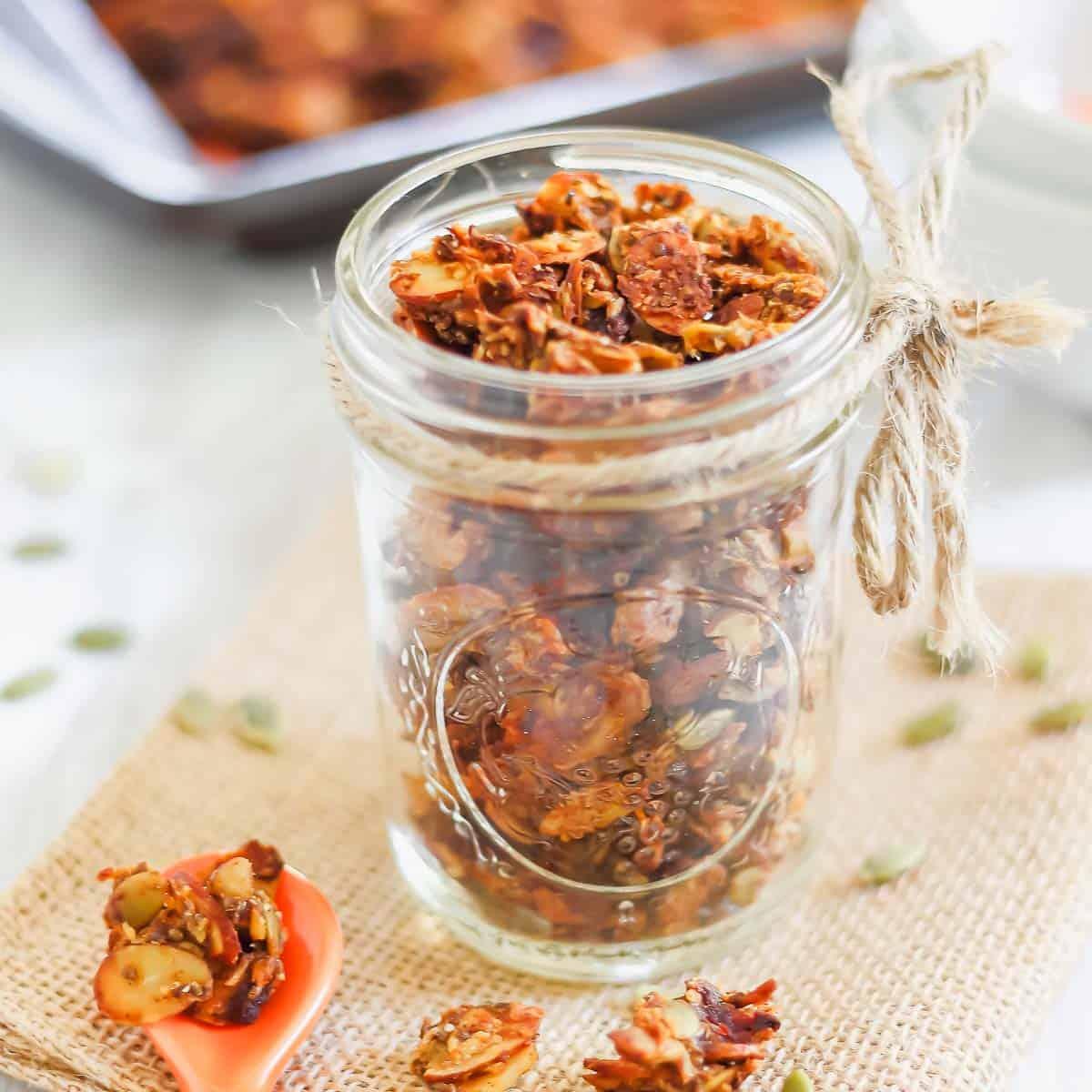 A clear glass jar filled with a homemade pumpkin granola recipe tied with a twine bow, sitting on a piece of burlap. A spoon with some granola is placed nearby, and a baking tray with more granola is in the background.