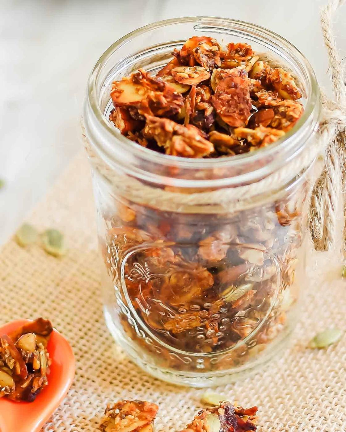A glass jar filled with granola, placed on a burlap cloth. Some granola pieces are scattered around the jar and in a small orange dish beside it.