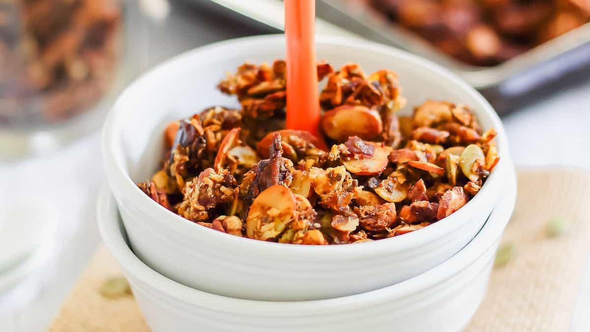 A white ceramic bowl filled with pumpkin granola and a red spoon placed in the middle. The granola contains visible nuts and seeds.