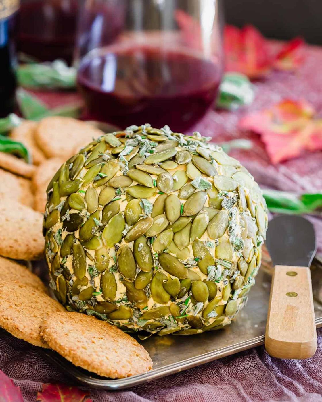 A large cheese ball coated in pumpkin seeds is displayed on a tray with crackers and a butter knife, with a glass of red wine in the background.