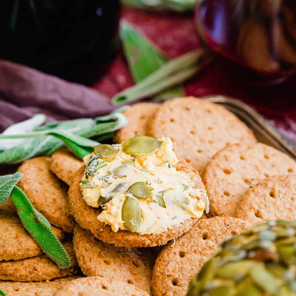 A pile of round, crispy biscuits, with one biscuit topped with a creamy pumpkin cheese spread and pumpkin seeds, garnished with leafy herbs. A maroon cloth and a glass container are in the background.