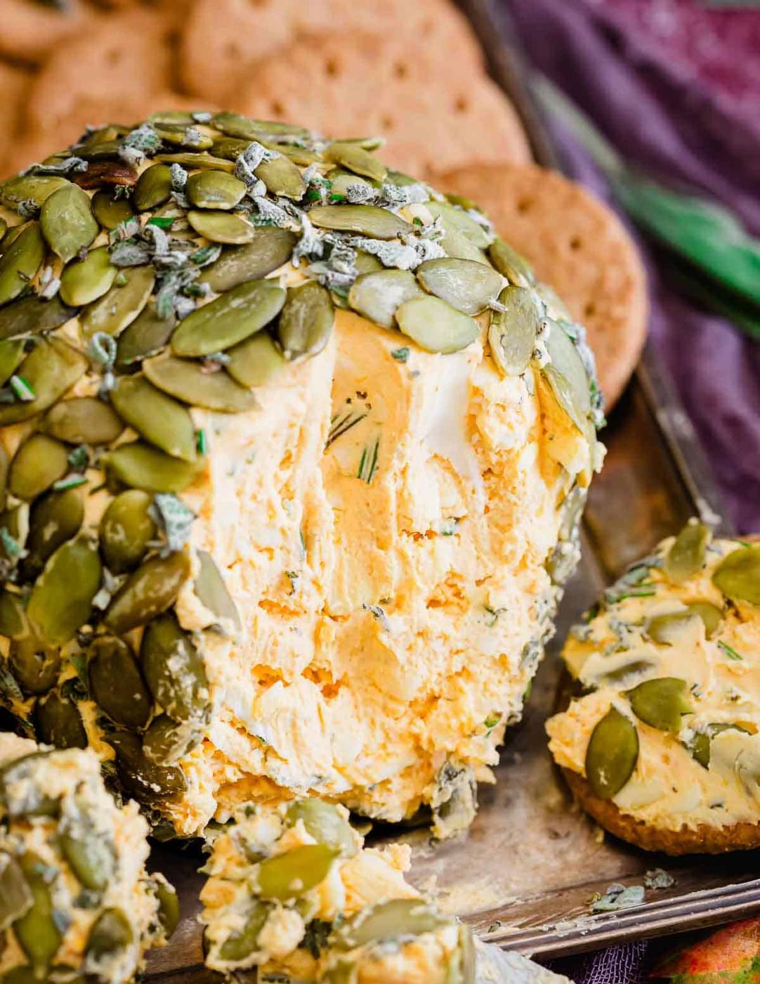 A close-up of a pumpkin cheese ball covered with pumpkin seeds with a chunk taken out, surrounded by crackers.