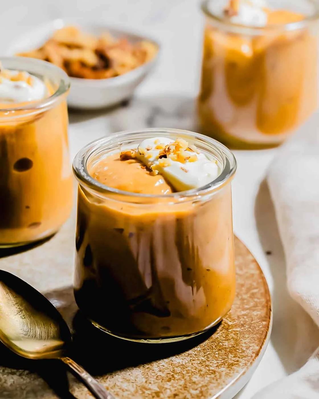 A close-up of glass jars filled with creamy pumpkin pudding, topped with whipped cream and a sprinkle of spices, set on a plate with a spoon and a bowl of crunchy toppings in the background.