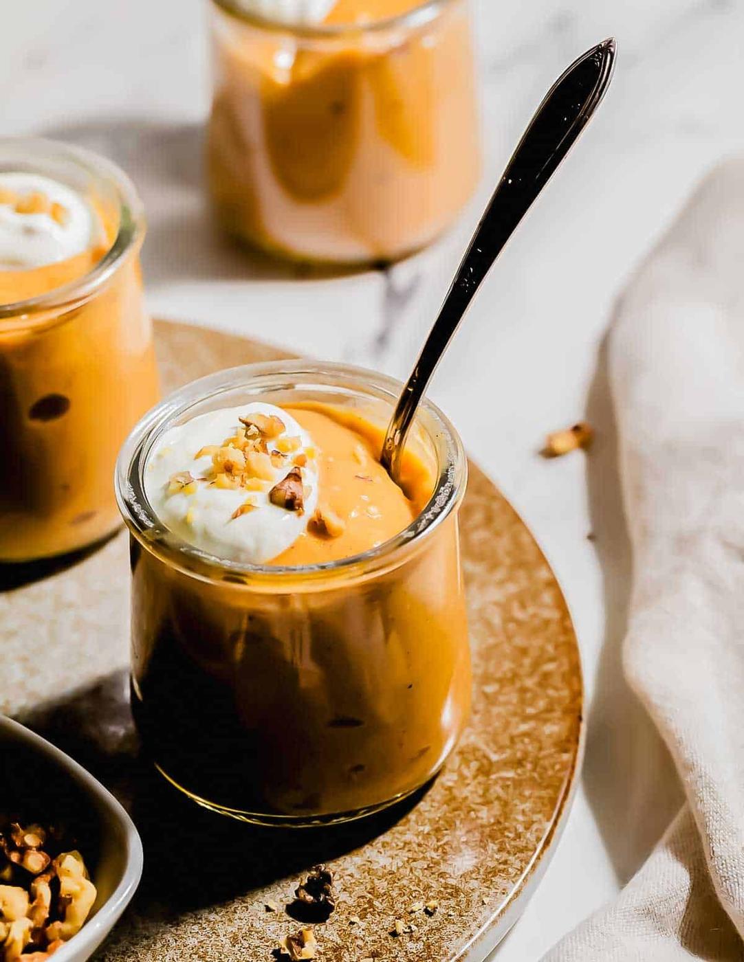 A small glass jar of pumpkin pudding topped with whipped cream and chopped nuts, with a spoon inside, sits on a round tray next to another jar in the background.