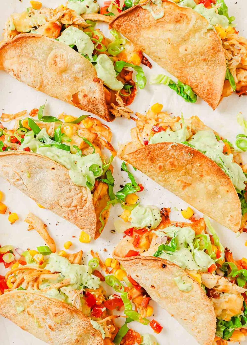 Six crispy tacos filled with shredded chicken, corn, diced tomatoes, lettuce, and topped with a drizzle of avocado crema are arranged on a white surface.