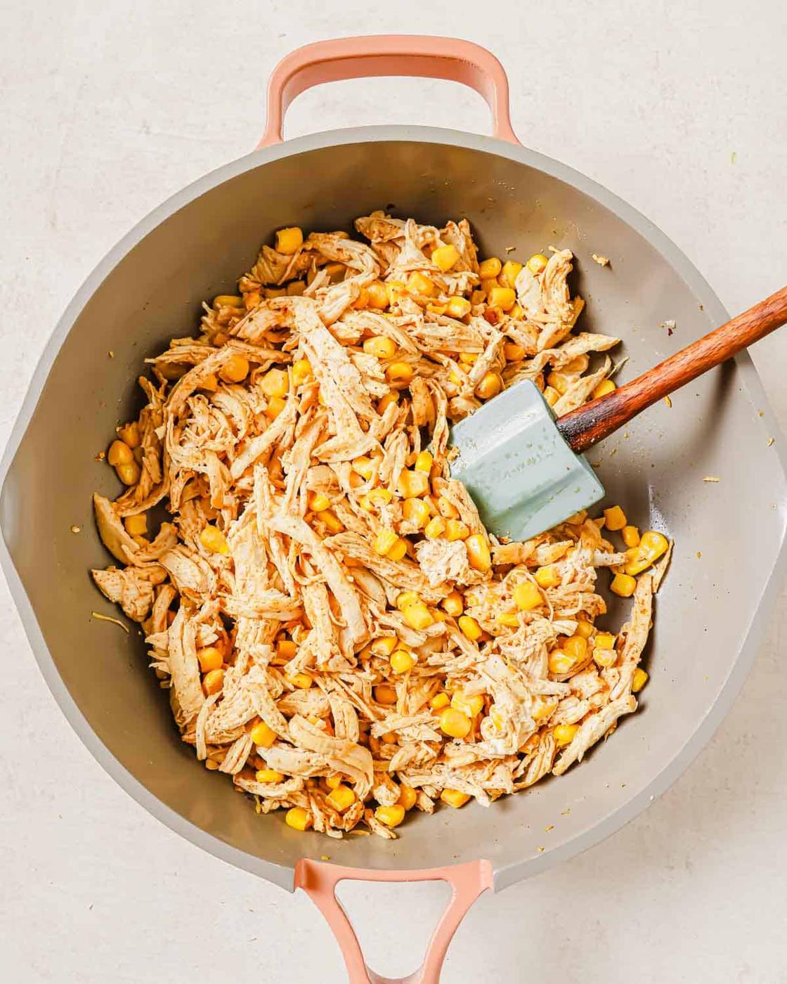 A skillet with shredded chicken and corn, mixed together and being stirred with a silicone spatula.