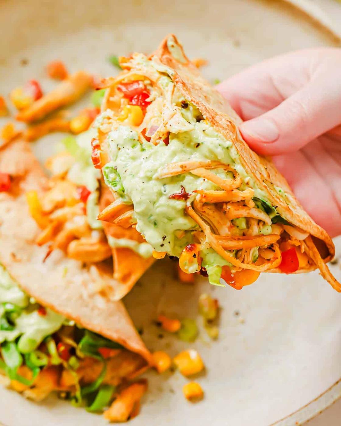 A hand holding a taco filled with fried chicken, lettuce, corn, diced tomatoes, and a creamy avocado sauce. Another taco on a plate is partially visible in the background.