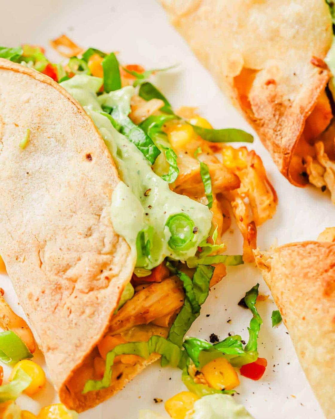 Close-up of a taco with chicken, diced vegetables, shredded lettuce, and green sauce, with another taco partially visible in the background.