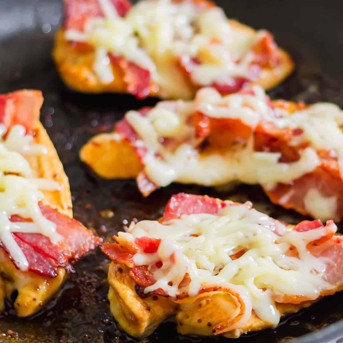 Close-up of four chicken pieces topped with melted cheddar cheese and crispy bacon, cooked on a dark skillet. The golden-brown chicken contrasts with the creamy cheese and red-brown bacon, creating an inviting and savory appearance.